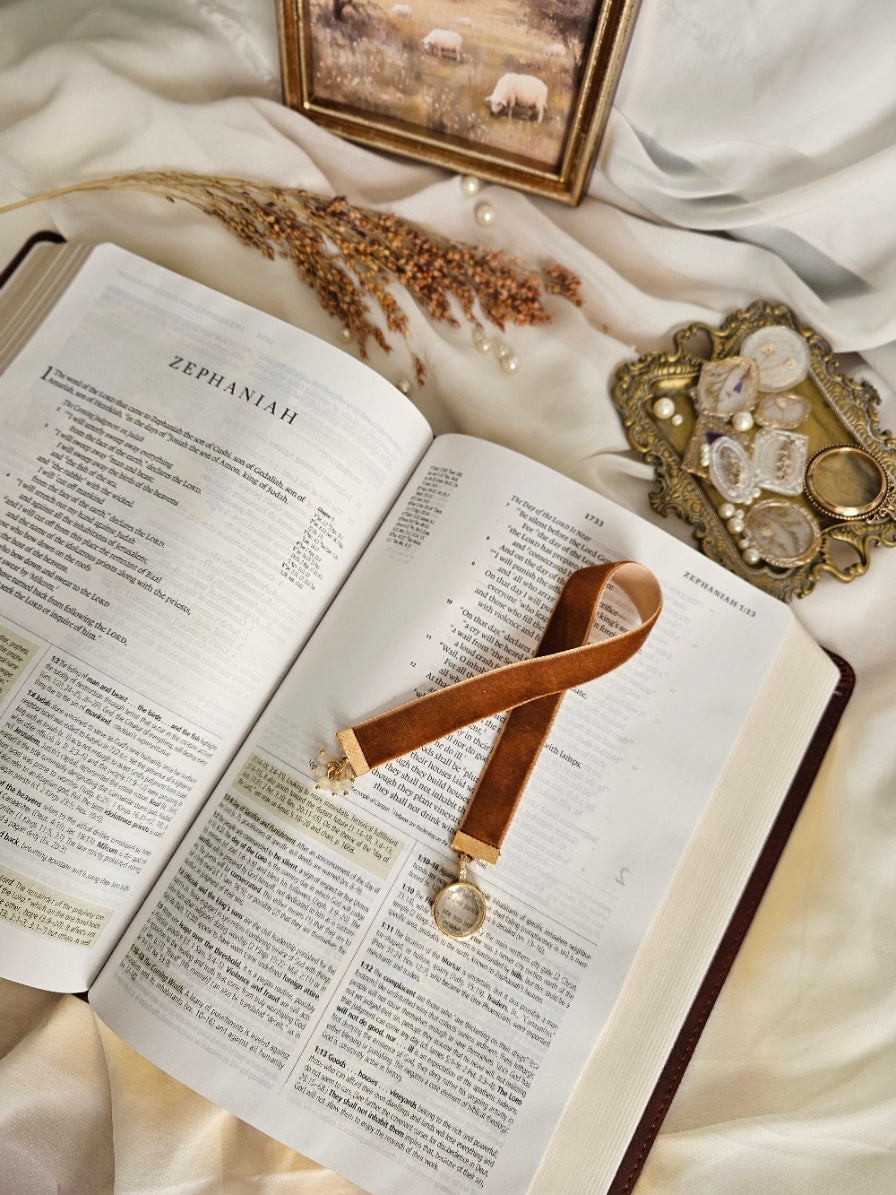 Caramel Velvet ribbon with gold locket charm on one end and cluster of beads charm on another end laying on top of the Bible. 