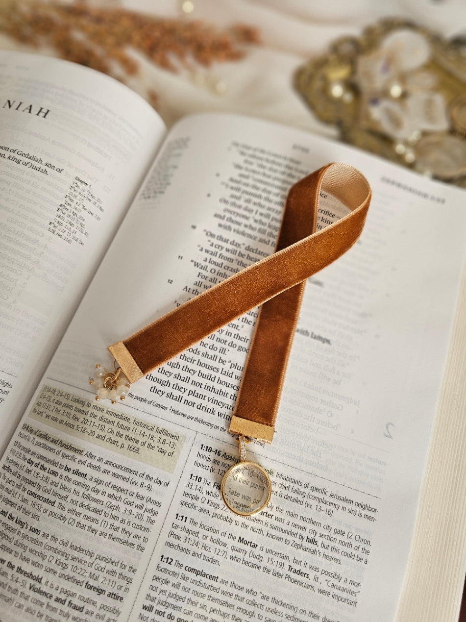 Caramel Velvet ribbon with gold locket charm on one end and cluster of beads charm on another end laying on top of the Bible. 