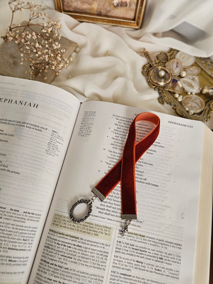 Burnt red-orange velvet ribbon with silver frame and cross charms on each end sitting on an open Bible.