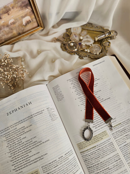Burnt red-orange velvet ribbon with silver frame and cross charms on each end sitting on an open Bible.