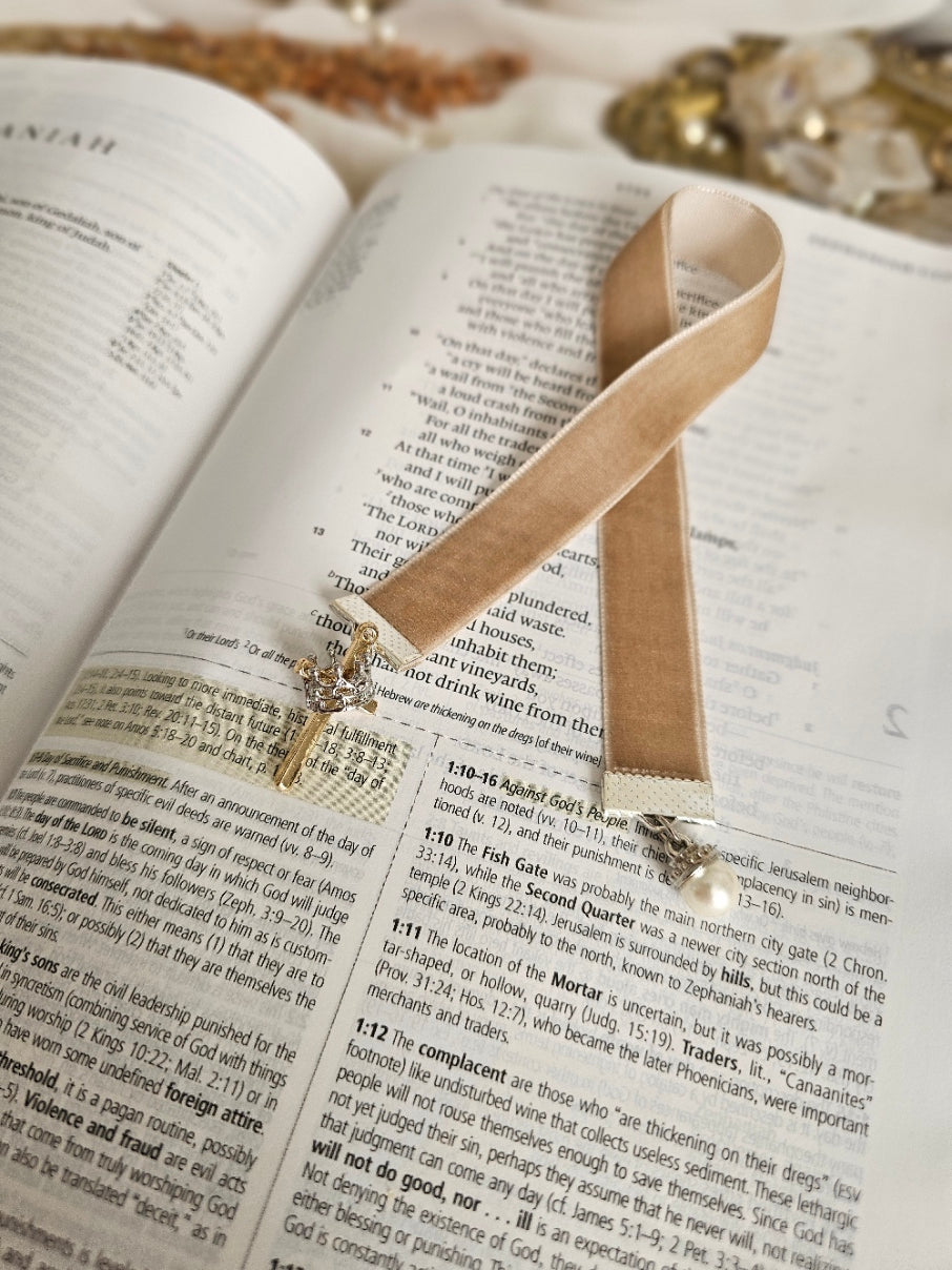 Tan Velvet ribbon with gold cross and silver crown charm on one end and pearl charm on another end laying on top of the Bible. 