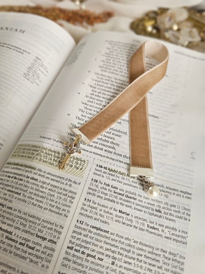 Tan Velvet ribbon with gold cross and silver crown charm on one end and pearl charm on another end laying on top of the Bible. 