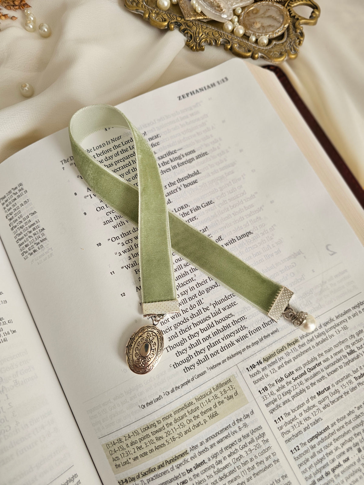 Sage velvet ribbon with silver locket and pearl charms on each end sitting on top of an open Bible. 