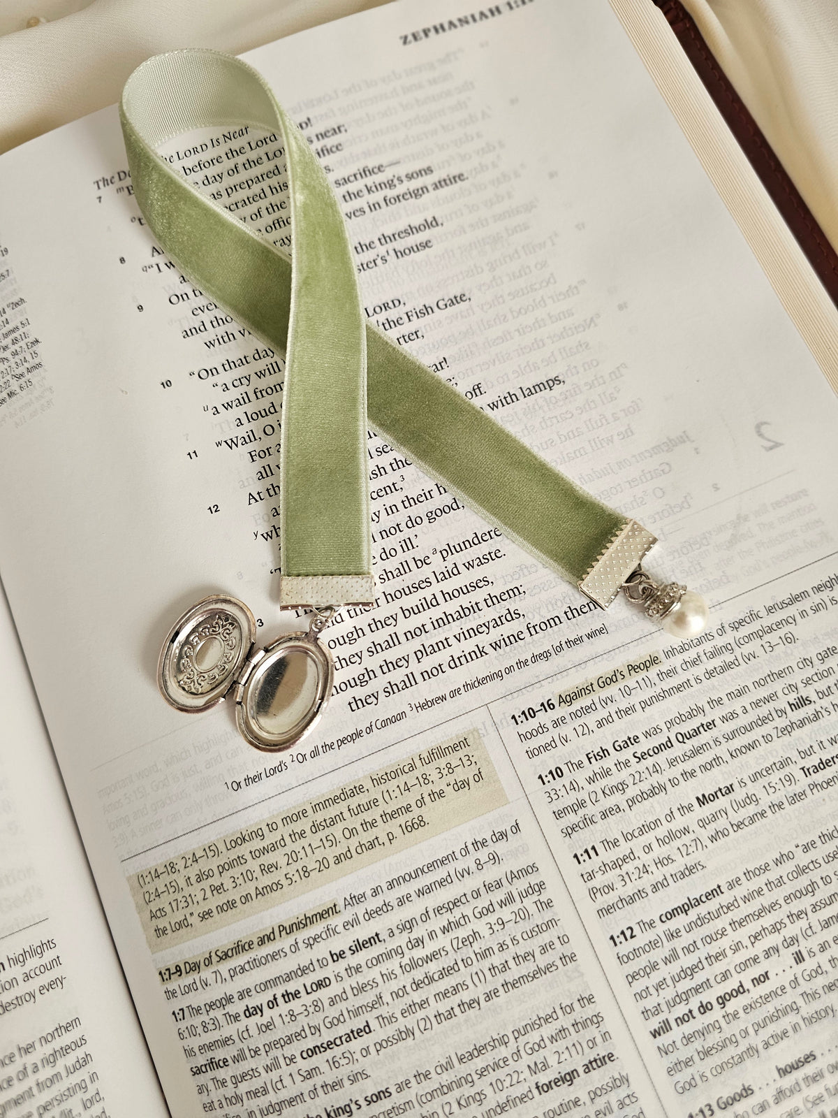 Sage velvet ribbon with open silver locket and pearl charms on each end sitting on top of an open Bible.