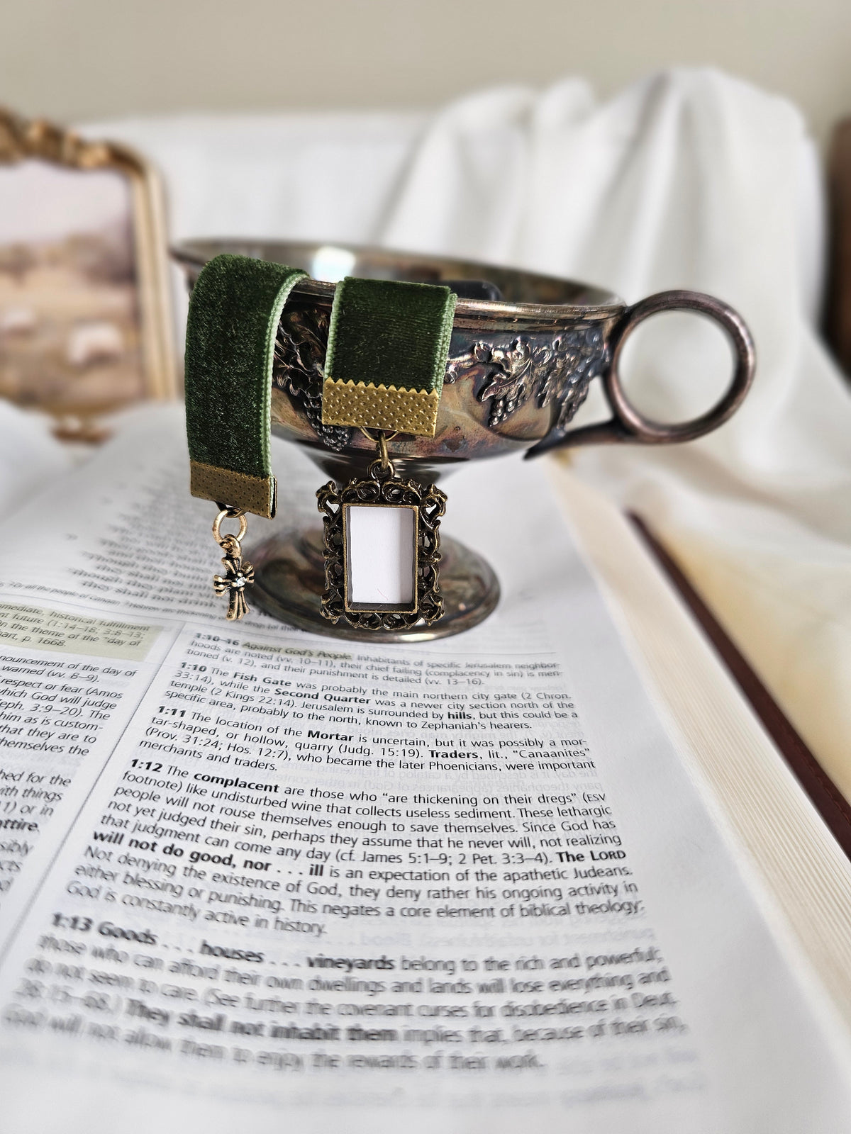 Dark green velvet ribbon with brass frame and cross charms on each end sitting on an open Bible inside a metal cup.