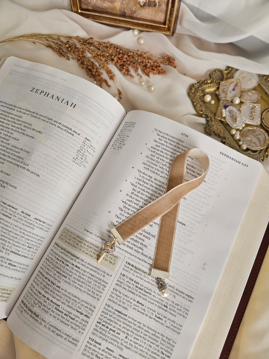 Tan Velvet ribbon with gold cross and silver crown charm on one end and pearl charm on another end laying on top of the Bible. 