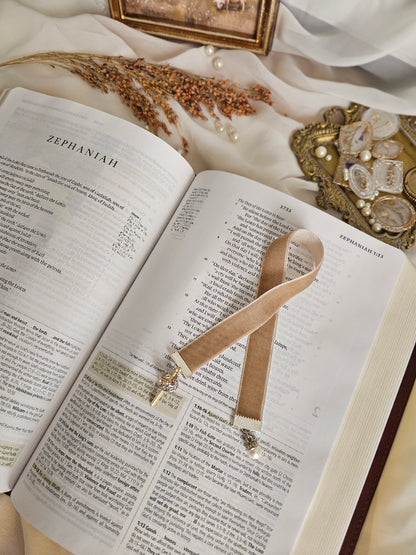 Tan Velvet ribbon with gold cross and silver crown charm on one end and pearl charm on another end laying on top of the Bible. 