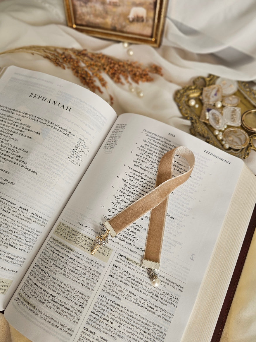 Tan Velvet ribbon with gold cross and silver crown charm on one end and pearl charm on another end laying on top of the Bible. 